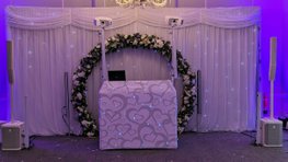 white dj booth with white lighting and white speakers in front of a white twinkle backdrop and flower arch