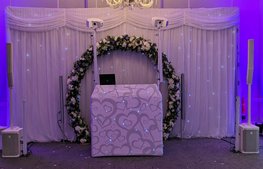 white dj booth with white lighting and white speakers in front of a white twinkle backdrop and flower arch